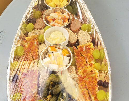 Charcuterie boat with assorted meats, cheeses, fruits, pickles, and olives arranged on a foil-wrapped tray with grapes and skewers