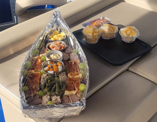 Charcuterie platter wrapped in foil on a boat bench, with meats, cheese, pickles, and fruit bowls nearby on a tray