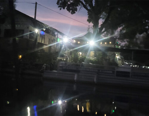 Night waterfront scene at dusk with a moored boat, illuminated buildings, and reflections in the water.