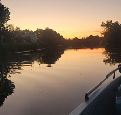 Sunset over a calm river seen from a boat, with silhouetted trees and reflections on the water.