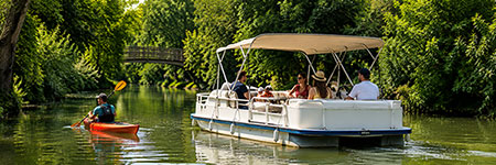 White pontoon boat with passengers under a canopy on a calm river, while a person in a red kayak paddles nearby among green riverbank trees.